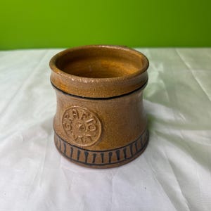 May include: A handcrafted, brown ceramic mug with a wide opening and a cylindrical shape. The mug features a decorative circular emblem and black painted accents. The mug is approximately 4 inches tall.