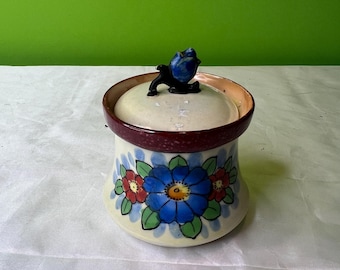 May include: A white ceramic jar with a floral design. The jar has a lid with a blue flower knob. The jar is decorated with blue, red, and green flowers.