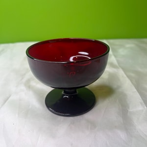 May include: A dark red glass goblet with a rounded bowl and a short, circular base. The glass has a glossy finish, and the interior of the bowl reflects light. The goblet is likely for serving beverages.