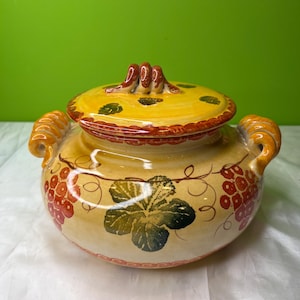 May include: A ceramic serving bowl with a lid, featuring a yellow and tan color scheme. The bowl is adorned with red grapes and green leaf designs. It has two handles and a decorative lid with a textured handle.