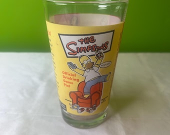 May include: A yellow glass pint glass with a cartoon image of Homer Simpson sitting in a chair with a remote control and a can of beer. The glass has the text "The Simpsons" and "Official Drinking Game Pint" printed on it.