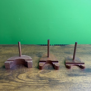 May include: Three brown wooden stands, each featuring a semi-circular base with a central vertical rod. The stands have a notched design on the front edge, providing a stable platform for holding items upright in a display or storage configuration.