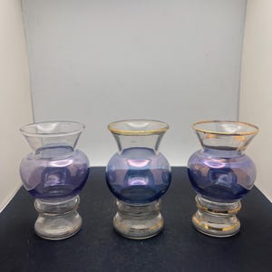 May include: Three small, decorative glass vases. Each vase has a clear base and a bulbous, iridescent purple body. The rims of two vases are trimmed with gold. The vases are designed with a unique shape, suitable for small floral arrangements or decorative display.