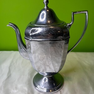 May include: A silver-colored teapot with a lid and handle. The teapot has a decorative floral pattern on the body and sits on a round base. The background is a solid green.