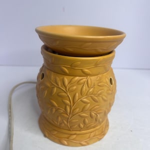 May include: Mustard yellow electric oil warmer with a leaf design. The warmer has a round dish on top and a base with a detailed embossed leaf pattern. The warmer has a power cord attached.