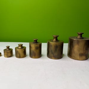 May include: A collection of six antique brass weights, each with a cylindrical body and a small handle. The weights are arranged in a row, increasing in size. They are displayed on a white surface against a green backdrop.