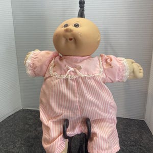 Könnte beinhalten: Eine Puppe mit einem runden, beigen Stoffkopf, Grübchen in den Wangen und braunen Augen. Sie trägt einen rosa-weiß gestreiften Strampler mit kurzen Ärmeln, Spitzenbesatz, einem weißen Spitzenkragen und einer kleinen Schleife am Ausschnitt.