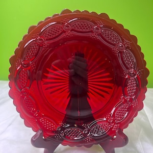 Könnte beinhalten: Eine rubinrote Glasplatte mit einem gewellten Rand und aufwendigen geprägten Details. Die Platte wird auf einem dunkelroten Ständer präsentiert, der ihre leuchtende Farbe und ihr Design hervorhebt. Das Zentrum der Platte zeigt ein Sonnenstrahlenmuster.