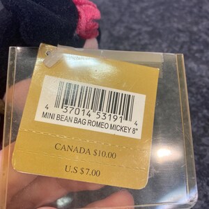 May include: A plush toy tag featuring a barcode and the text MINI BEAN BAG ROMEO MICKEY 8". The tag is encased in a clear plastic protector and displays pricing for Canada and the U.S. The plush toy is partially visible with black and pink fabric.