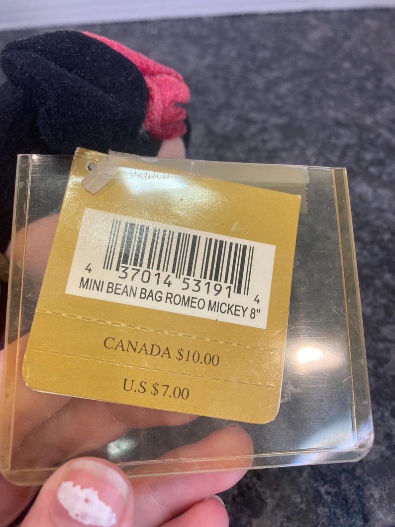 May include: A plush toy tag featuring a barcode and the text MINI BEAN BAG ROMEO MICKEY 8". The tag is encased in a clear plastic protector and displays pricing for Canada and the U.S. The plush toy is partially visible with black and pink fabric.