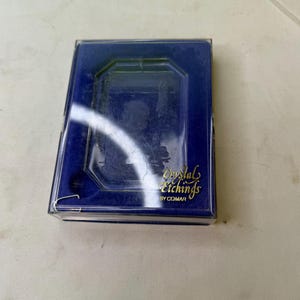 May include: A rectangular, navy blue box with a clear plastic lid. The box contains a crystal etching. The text "Crystal Etchings by COMAR" is printed in gold script on the box.