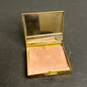 May include: A gold-colored compact mirror with a hinged lid and a pink velvet interior. The mirror is open and the lid is propped up.