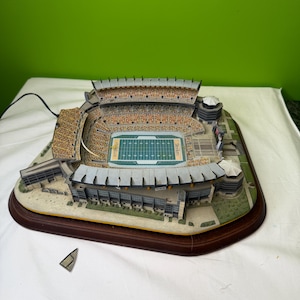 May include: A detailed miniature stadium model with a green field and seating areas. The model is set on a dark wooden base and features the word "Saints" on the field. The stadium has a detailed exterior and is lit with a cord.