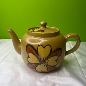 May include: A ceramic teapot with a lid, featuring a floral design in yellow, orange, and brown. The teapot is a muted olive green color. The handle and spout are curved. The teapot is sitting on a white surface.