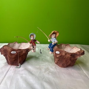 Może przedstawiać: Dwie ceramiczne donice w kształcie pni drzew, każda z małą figurką dziecka łowiącego ryby. Jedno dziecko ma na sobie czerwoną sukienkę w kropki, drugie koszulę w niebiesko-białe paski. Donice są brązowe z białym wnętrzem.