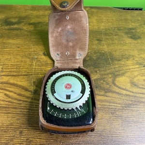 May include: Vintage light meter in a brown leather case. The meter has a circular dial with numbers and markings, and a small red logo. The case is open, revealing the meter's face. The case has a flap that fastens with a snap.