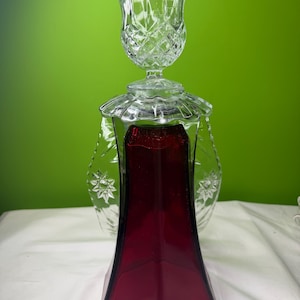 May include: A clear glass candle holder with a ruby red base and a tulip-shaped top. The base is a geometric shape, and the top has a diamond pattern. The candle holder is on a white surface against a green background.
