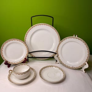 Op de afbeelding: Een verzameling witte porseleinen borden met een goud-bruine bloemenrand. De set bevat borden van verschillende formaten, een kop en schotel en een klein bord. De borden worden gepresenteerd op standaards en een zwart metalen rek tegen een groene achtergrond.