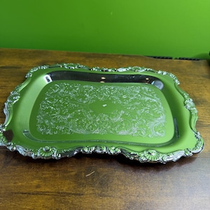 May include: A rectangular, silver-coloured serving tray with a green interior. The tray has ornate, scrolling edges and a detailed, embossed floral pattern in the centre. The tray is displayed on a wooden surface.