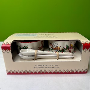以下が含まれることがあります： セラミック製のスプーンとトレイが付いた調味料ポットセット。セットには、お祝いのイラストが描かれた2つの小さな白いセラミックポットと、お揃いのスプーンが2つ含まれています。パッケージは、赤と白の装飾トリムが付いたベージュ色の箱です。「Condiment Pot Set」の文字が箱に印刷されています。