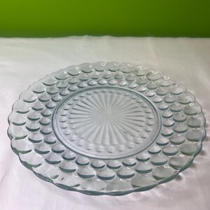May include: A clear glass plate with a raised, textured design. The plate has a scalloped edge and a centre with a sunburst pattern.