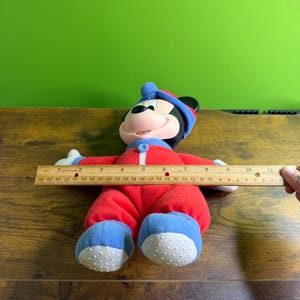 May include: A plush Mickey Mouse doll wearing a red onesie with blue accents and a matching hat. The doll is approximately 10 inches long. The doll's feet have white, textured soles. The doll is lying on a wooden surface.