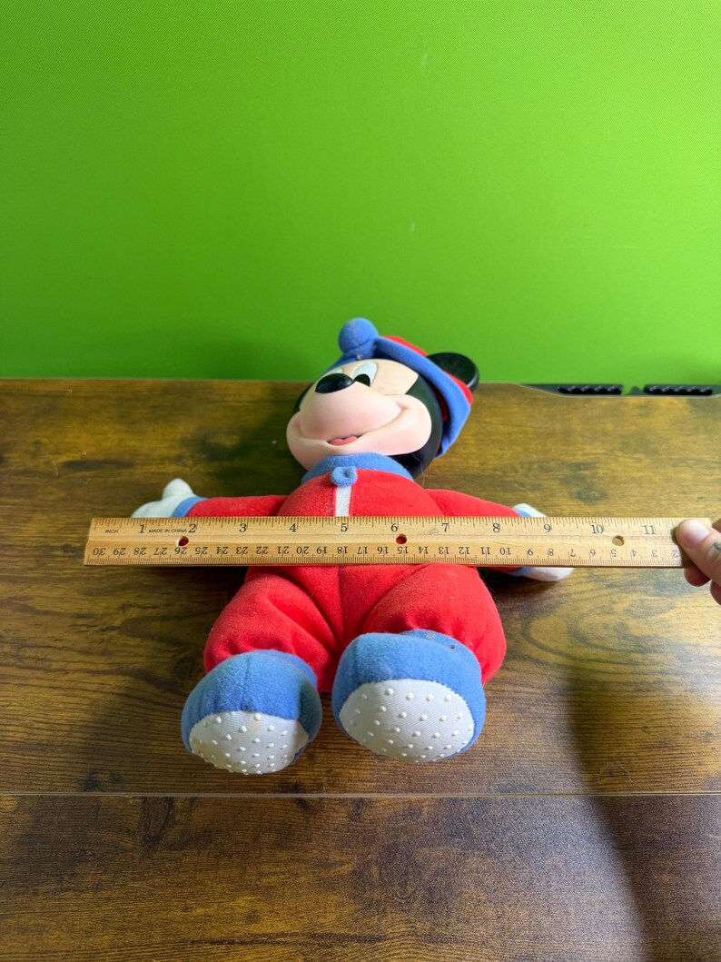 May include: A plush Mickey Mouse doll wearing a red onesie with blue accents and a matching hat. The doll is approximately 10 inches long. The doll's feet have white, textured soles. The doll is lying on a wooden surface.