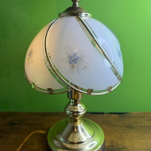 Peut inclure: Une lampe de table vintage en laiton avec un abat-jour blanc à motifs floraux. L'abat-jour a un design incurvé avec des bordures dorées et de petits accents en forme de cœur. Le pied de la lampe est rond et également en laiton.