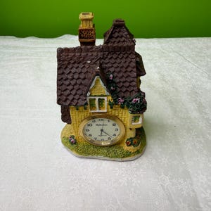 May include: A miniature house-shaped clock with a yellow facade, brown roof, and a chimney. The clock face is gold-toned with black numerals and hands. Decorative flowers and greenery adorn the house. The clock is a charming decorative piece.