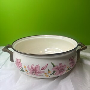 May include: Off-white enamel cooking pot with pink floral design. The pot has two bronze-colored handles and a bronze-colored rim. The floral design includes pink lilies and small yellow flowers. The pot is sitting on a white surface against a green background.