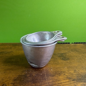 Può includere: Un set di tre misurini vintage in metallo, impilati. Ogni tazza ha una forma a ciotola arrotondata e un piccolo manico con un foro. Lo sfondo è verde pieno e le tazze sono su una superficie di legno.