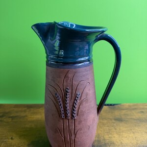 May include: A ceramic pitcher featuring a dark teal glazed upper section and a matte brown lower body. The brown portion is decorated with a carved wheat stalk motif. The pitcher has a curved handle and a wide, shaped pouring spout at the top.