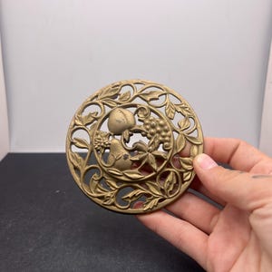 May include: A circular, antique brass decorative piece featuring a fruit and leaf design. The intricate openwork design includes a peach, grapes, and foliage. The piece is likely a decorative element for furniture or a similar item.