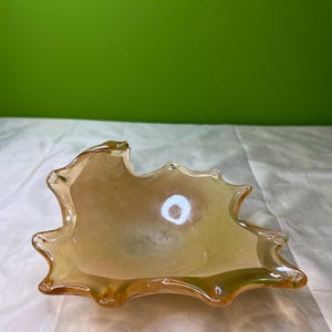 Peut inclure: Un bol décoratif en verre ambré, de forme irrégulière rappelant une feuille. Le bol a une finition brillante et un aspect légèrement irisé. Les bords sont inégaux, ce qui ajoute à son design unique. Le bol est vide et posé sur une surface blanche.