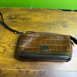 May include: A brown and green leather wristlet with a crocodile pattern. The wristlet has a zipper closure and a small green label. A black wrist strap is attached to the side.