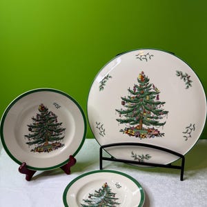 May include: A set of three Christmas-themed plates with a green border. Each plate features a detailed illustration of a decorated Christmas tree. The plates are displayed on a white surface with a green background.