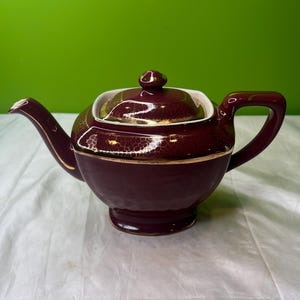 Op de afbeelding: Een bordeauxrode keramische theepot met bijpassende deksel. De theepot heeft gouden randen en accenten. De handgreep en de tuit zijn gebogen. De theepot staat op een wit oppervlak met een groene achtergrond.
