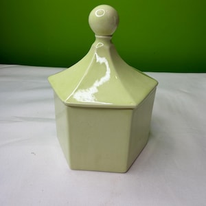 Pode incluir: Um recipiente de cerâmica verde pálido com uma base hexagonal e uma tampa em estilo pagoda. A tampa é encimada por um botão redondo. O recipiente está sobre um fundo verde e uma superfície branca.