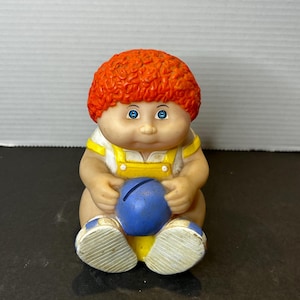 May include: A vintage Cabbage Patch Kid piggy bank with red curly hair, blue eyes, and a yellow and blue outfit. The piggy bank is holding a blue ball with a slot for coins.