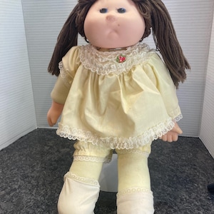 Könnte beinhalten: Eine Puppe mit braunen Wollhaaren, blauen Augen und Sommersprossen, bekleidet mit einem hellgelben Kleid mit Spitzenkragen, einem kleinen Blumenakzent, passender gelber Pumphose und weißen Stoffschuhen mit einem weichen, runden Gesicht.