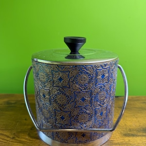 Peut inclure: Un seau à glace cylindrique présentant un extérieur à motifs bleus et dorés avec une finition métallique. Le récipient comprend une anse pivotante en métal argenté, un couvercle argenté assorti et un bouton noir centré sur le couvercle.