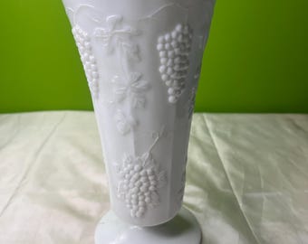 May include: White glass vase with a flared top and a pedestal base. The vase features a raised grape and leaf design around the body. The vase is approximately 25 cm tall.