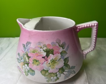 May include: A vintage pink and white ceramic jug with a floral design. The jug has a handle and a spout, and the floral design features pink, white, and green flowers. The jug has a crack near the spout.