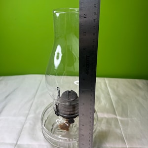 Puede incluir: Una l&aacute;mpara de aceite de vidrio transparente con base redonda y chimenea. La l&aacute;mpara tiene un ajustador de mecha de metal y una chimenea de vidrio transparente. La regla indica una altura de unos 30 cm.