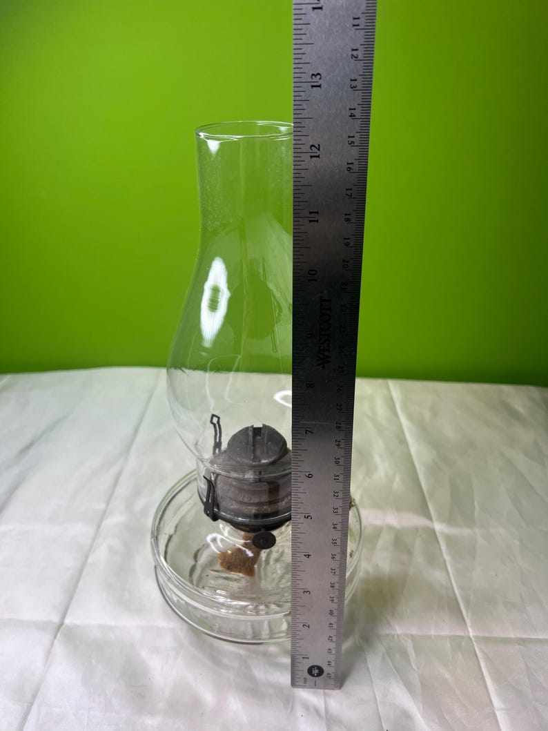 Puede incluir: Una l&aacute;mpara de aceite de vidrio transparente con base redonda y chimenea. La l&aacute;mpara tiene un ajustador de mecha de metal y una chimenea de vidrio transparente. La regla indica una altura de unos 30 cm.