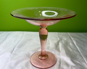 May include: A pink glass pedestal dish with a round base, a slender stem, and a wide, shallow bowl. The dish is made of transparent glass and sits on a white surface against a green background. The dish is likely used for serving desserts or displaying decorative items.
