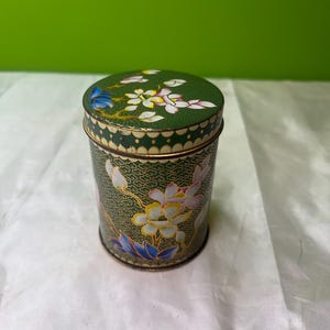 Pode incluir: Uma lata cilíndrica vintage com tampa. A lata é verde escuro com um desenho floral em branco, amarelo e azul. A tampa e o corpo têm um padrão repetitivo. A lata é provavelmente usada para armazenamento.