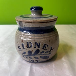 Puede incluir: Un pequeño tarro redondo de cerámica con tapa. El tarro está decorado con rayas azules y blancas y un diseño floral azul. El tarro dice "Sidney NY" en la parte delantera.