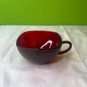 May include: A deep red glass teacup with a rounded square shape and a small handle. The interior of the cup is a brighter red, contrasting with the darker exterior. The cup sits on a white surface against a green background.
