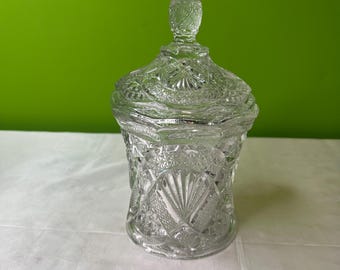 May include: A clear, cut-glass jar with a lid. The jar has a detailed, textured pattern with a knob on top of the lid. The jar is sitting on a white surface with a green background.